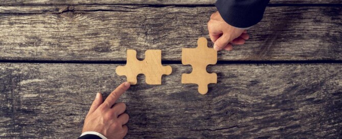 Zwei Hände in Anzügen halten zwei große Holz‑Puzzleteile, die kurz vor dem Zusammenfügen stehen.
